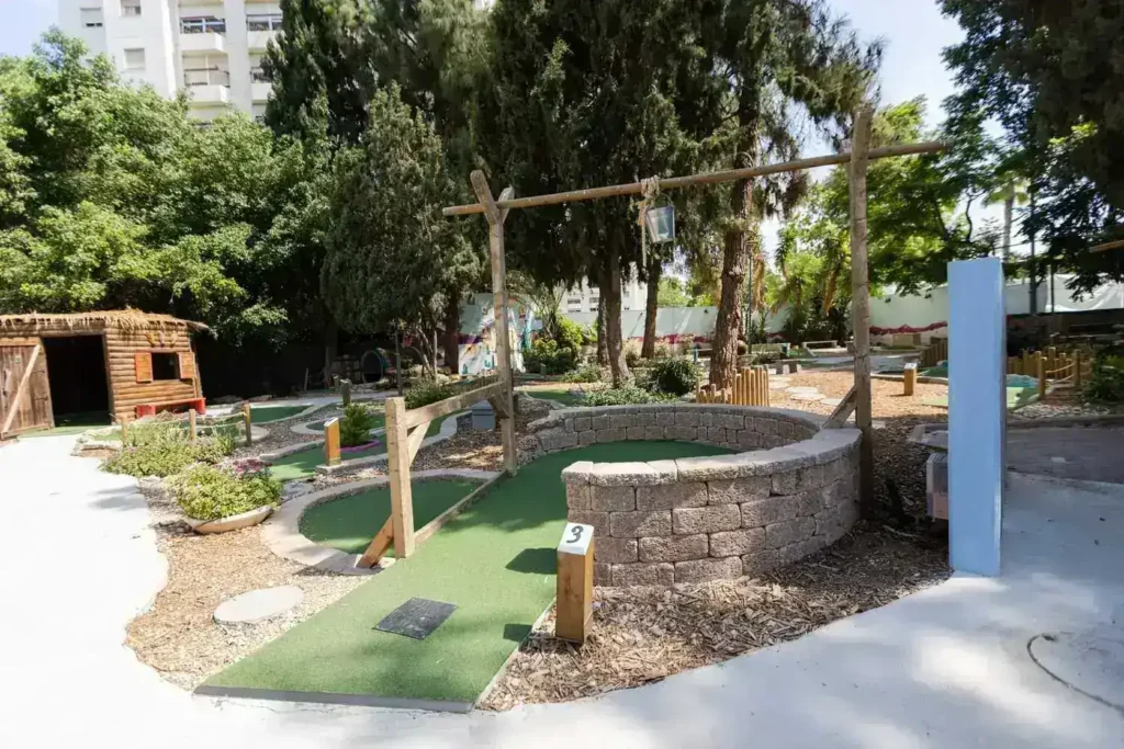 מגרש מיני גולף עם מסלול ירוק, עצים מסביב ופינות ישיבה, מתאים לפעילות משפחתית מגבשת.